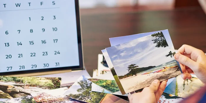 Eine Person schaut sich viele ausgedruckte Fotos am Schreibtisch an, daneben steht ein Laptop mit Kalender