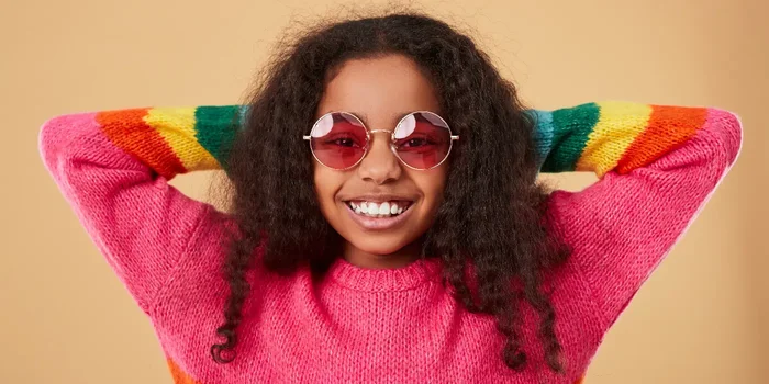 Ein lachendes Mädchen trägt einen pinken Pulli mit Regenbogenstreifen und eine rosa Sonnenbrille