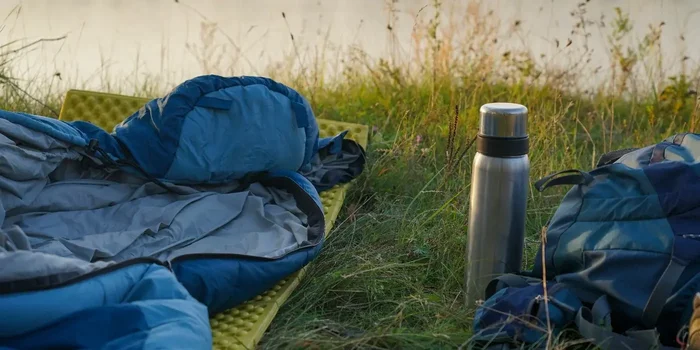 Schlafsack und Isomatte liegen neben Thermoskanne und Rucksack an einem Seeufer