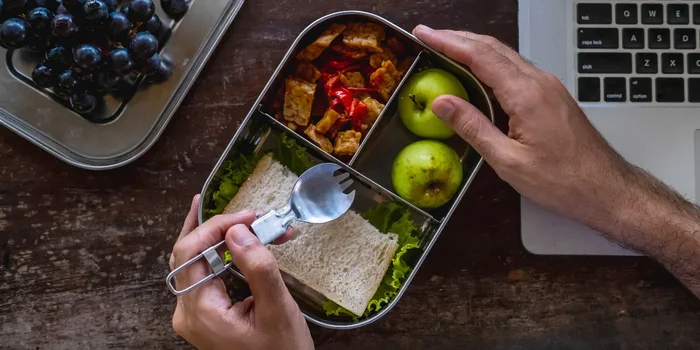 Zwei Hände halten eine rechteckige Edelstahl-Brotdose, die mit Brot, Snacks und Obst befüllt neben einem Laptop auf dem Tisch steht