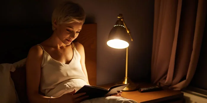 Eine Frau sitzt abends beim Licht einer Tischlampe im Bett und liest ein Buch