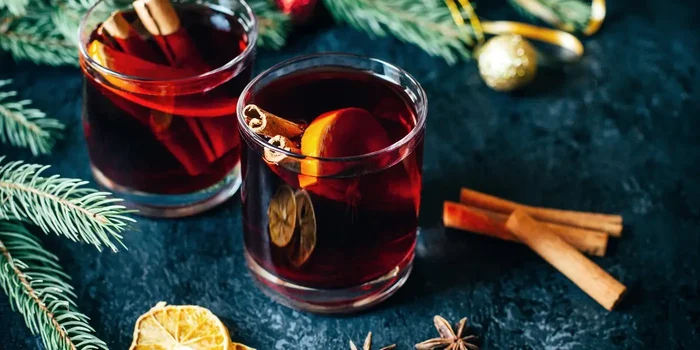 Zwei Gläser mit rotem Glühwein sowie Nelken, Zimtstangen und Orangenhälften befinden sich vor einem weihnachtlich dekorierten Hintergrund