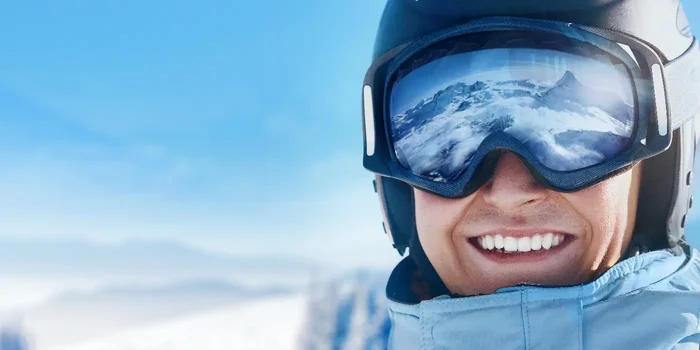 Ein lächelnder Mann mit schwarzem Helm, hellblauer Jacke und einer Schneebrille, in der sich die sonnige Berglandschaft spiegelt
