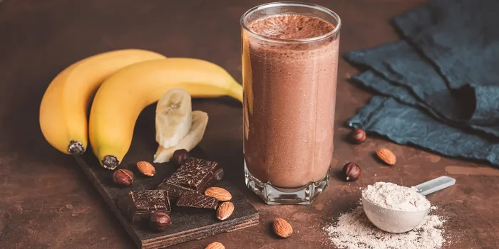 Ein Glas mit Schoko-Proteinshake steht auf einer braunen Oberfläche neben Schokolade, Nüssen, Bananen und einem Messlöffel Proteinpulver