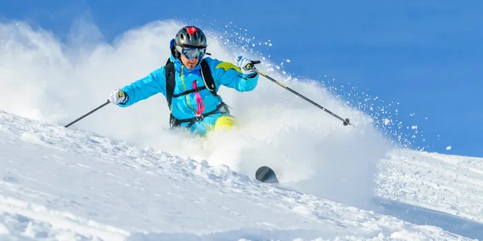 Ein Skifahrer in blauer Jacke fährt tief in den Schnee hinein, der dadurch aufgewirbelt wird