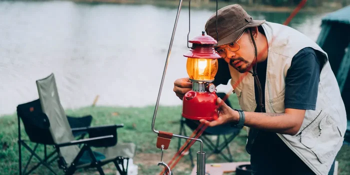 Ein Camper mit Hut und Weste beugt sich zu einer roten Petroleumlampe herab, um das Licht einzustellen