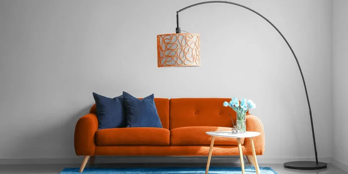 Eine orange Bogenlampe mit zylindrischem Textilschirm ist neben einem orangen Sofa mit türkisem Teppich und kleinem Beistelltisch positioniert