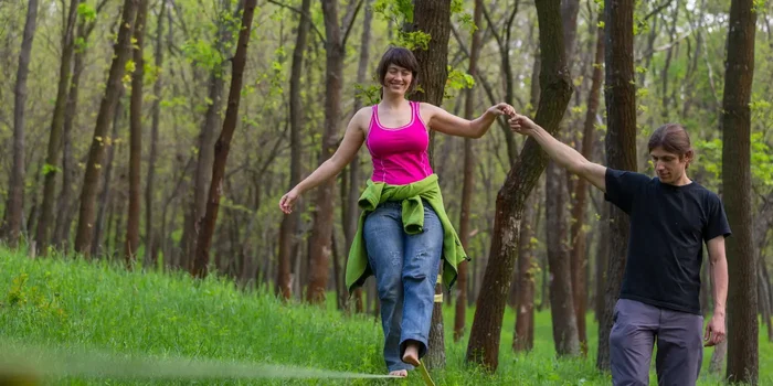 Eine lächelnde Frau balanciert mit einem Helfer an der Hand auf einer Slackline im Wald