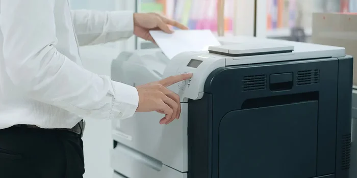Ein Mann, mit Hemd und Anzughose bekleidet, steht an einem Drucker und legt Papier in das Einzugsfach