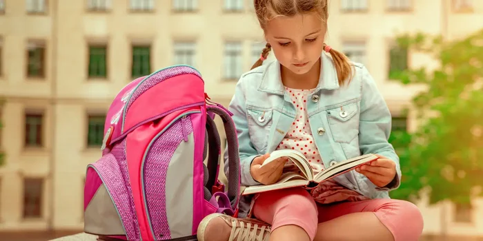 Ein junges Mädchen sitzt neben ihrem Schulranzen auf einem Tisch und liest in einem Buch