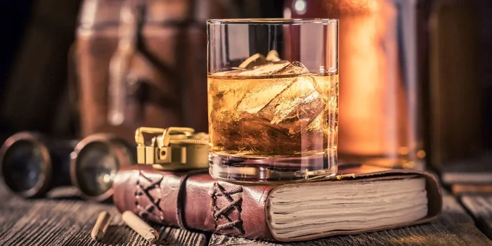 Ein Whiskyglas steht auf einem Buch, im Hintergrund ist eine Whiskyflasche