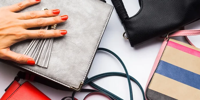 Eine Hand mit rot lackierten Nägeln liegt auf einer silbernen Handtasche, daneben liegen weitere Taschen