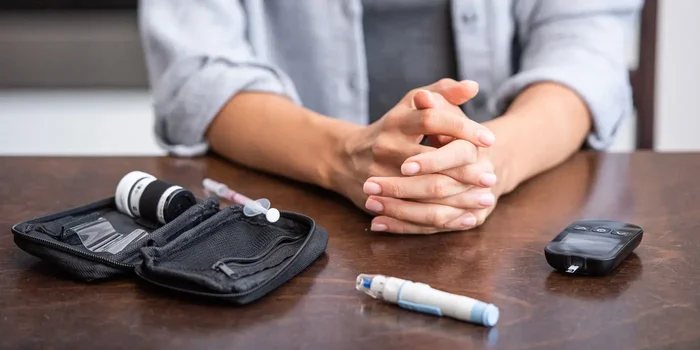 Auf einem dunklen Tisch liegt ein Etui mit Spritze, Insulin, Pen und Blutzuckermessgerät, im Hintergrund sitzt eine Person mit gefalteten Händen