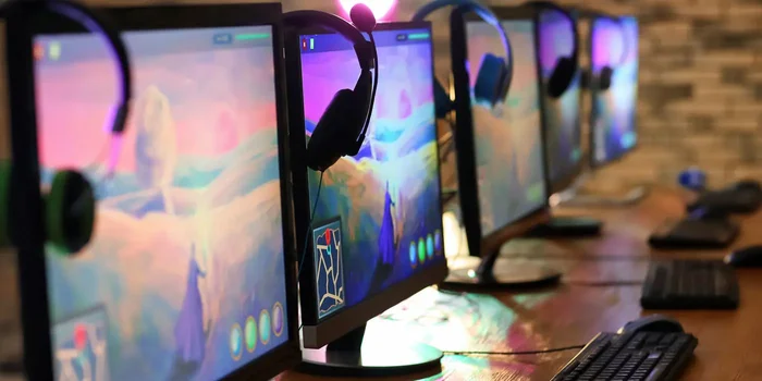 Mehrere Gaming Monitore befinden sich zusammen mit Kopfhörern, Tastaturen und PC-Mäusen auf Holztischen