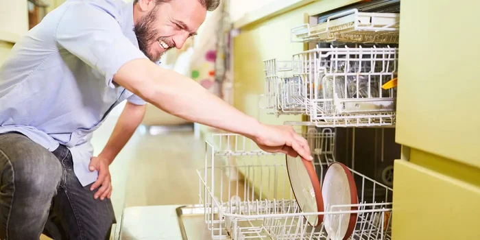 Mann kniet lachend vor Spülmaschine und räumt einen Teller in den unteren Korb