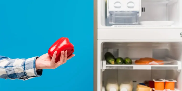 Hand, die eine rote Paprika hält neben einem geöffneten Kühlschrank mit Gefrierfach, in dem sich weitere Lebensmittel befinden