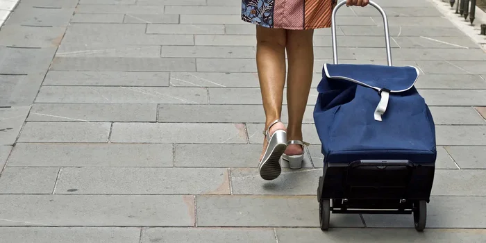 Eine Frau geht auf einem Fußgängerweg und zieht einen blauen Einkaufstrolley hinter sich her