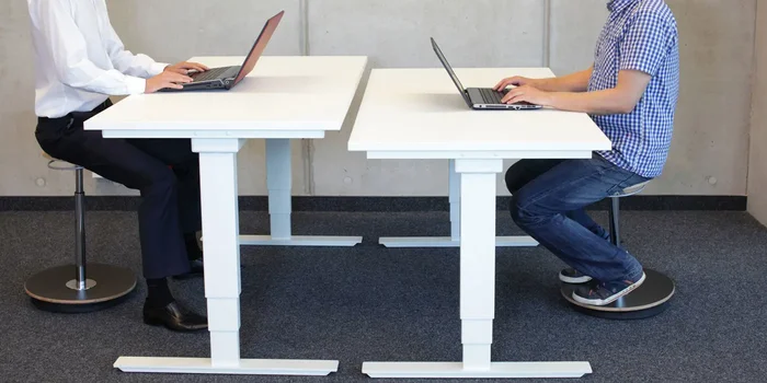 Zwei Männer sitzen sich an jeweils einem weißen, höhenverstellbaren Schreibtisch gegenüber und arbeiten an ihrem Laptop