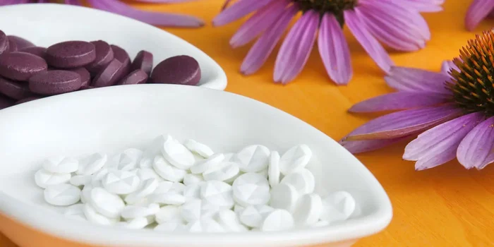 Tabletten liegen in Schüsseln auf einem hellen Holztisch, im Hintergrund lilafarbene Blüten