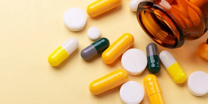 Vor einem gelben Hintergrund befindet sich eine braunes Tablettenglas mit verschiedenen ausgeschütteten Tabletten
