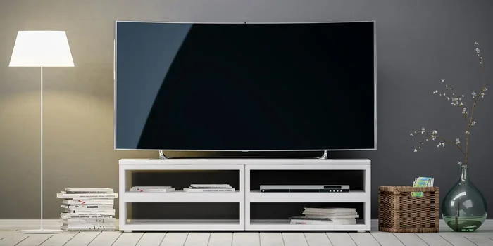 Ein Fernsehgerät steht vor einer grauen Wand auf einem kleinen TV-Schrank neben einer Stehlampe, einem Bücherstapel, einer Holzkiste und einer Glasvase